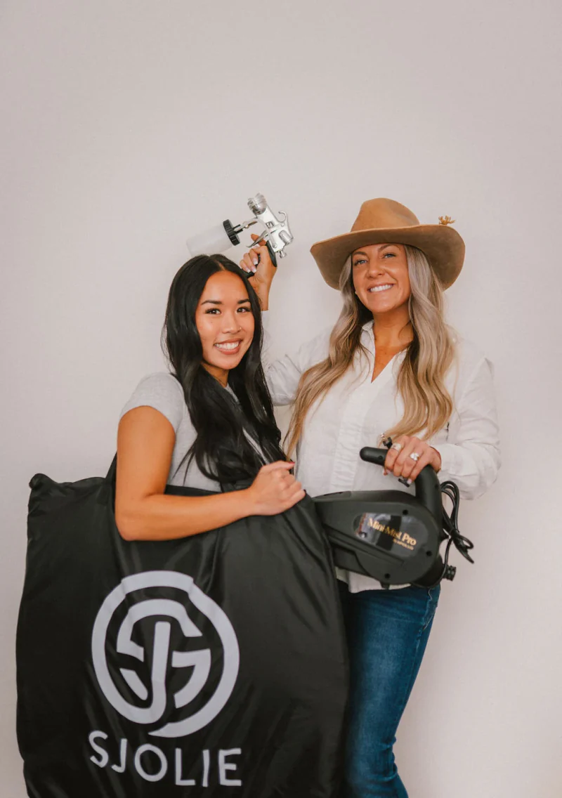 Two Sjolie spray tan students holding applicators, a turbine, and equipment bags